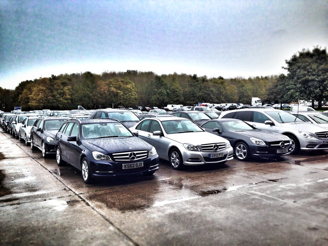Flera rader med Mercedes-Benz bilar uppställda på en auktionsplats utomhus med våt asfalt.